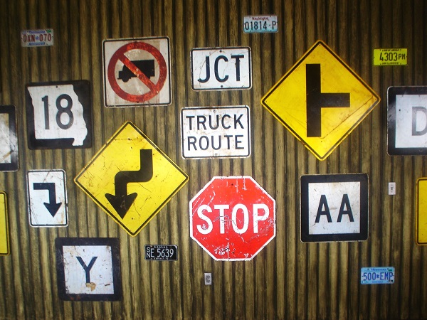 Assorted traffic signs, including stop, truck route, directional arrows, and road warnings, mounted on a weathered metal wall along with various license plates.