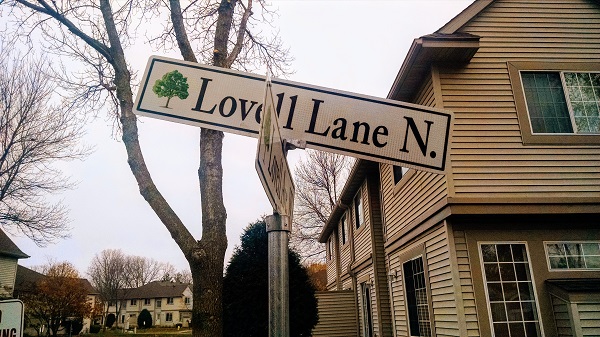 A bent and partially obscured street sign reading 'Lovell Lane N.' with a green tree symbol, mounted on a pole in a residential neighborhood with houses and bare trees in the background.